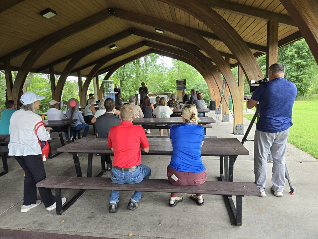 A group of people sitting on benches, listening to a speaker from the city at the Golden Lake delisting celebration event