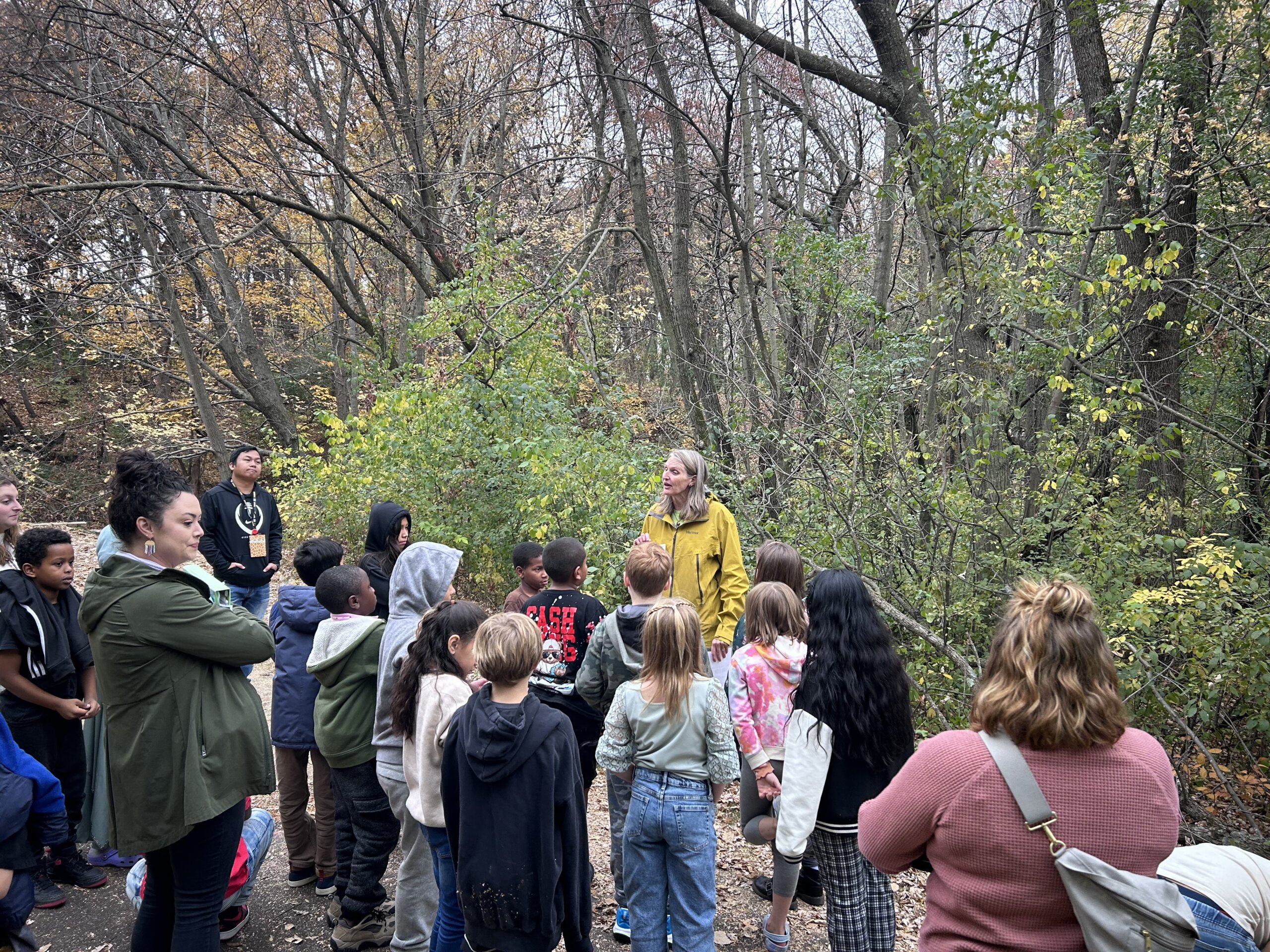 a group of students in front of teacher outside at a creek listening to a presentation about water quality. 